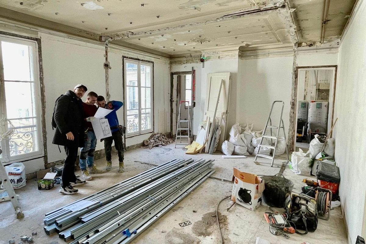 Séjour avant travaux : plafonds décroûtés, murs mis à nu, équipes du chantier consultant un plan.