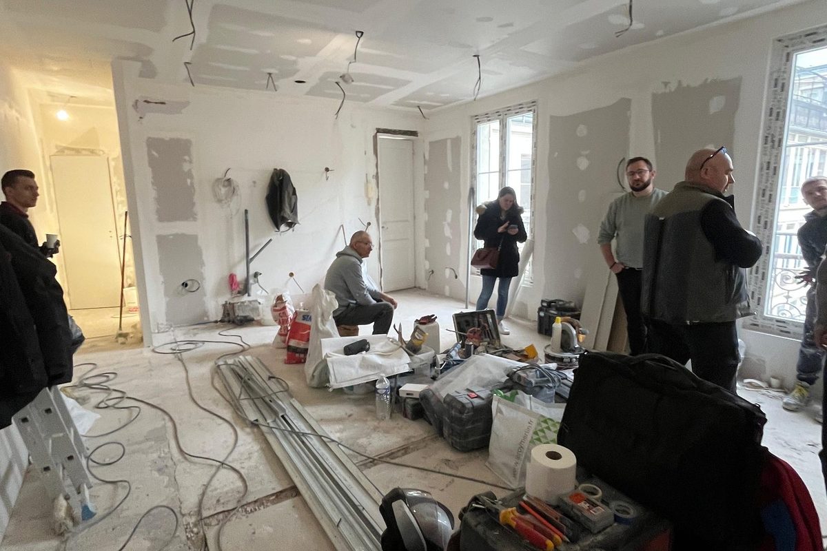 Chantier en cours : cloisons montées et enduites, outillage et gaines au sol, équipes en visite de chantier.